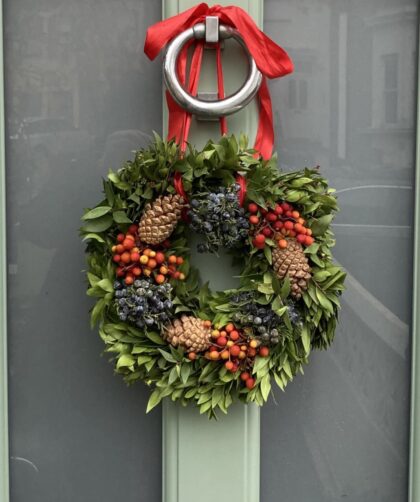 Photo of a Chriistmas wreate with green foliage, red berries and pine cones hanging from a knocker on a pale green door