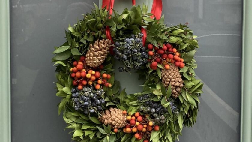 Photo of a Chriistmas wreate with green foliage, red berries and pine cones hanging from a knocker on a pale green door