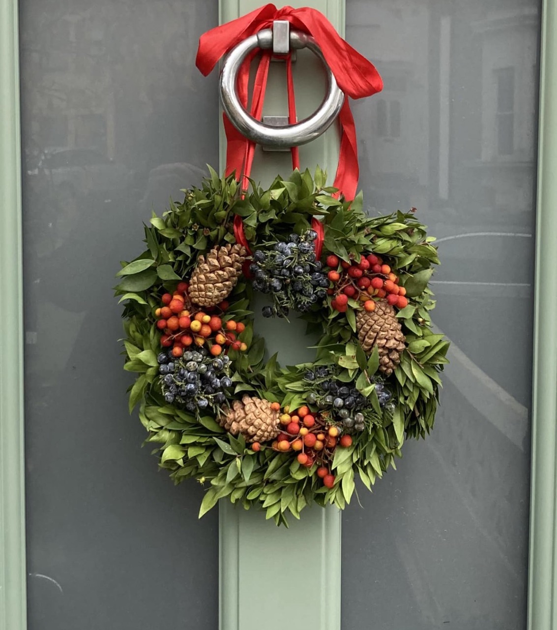 Photo of a Chriistmas wreate with green foliage, red berries and pine cones hanging from a knocker on a pale green door
